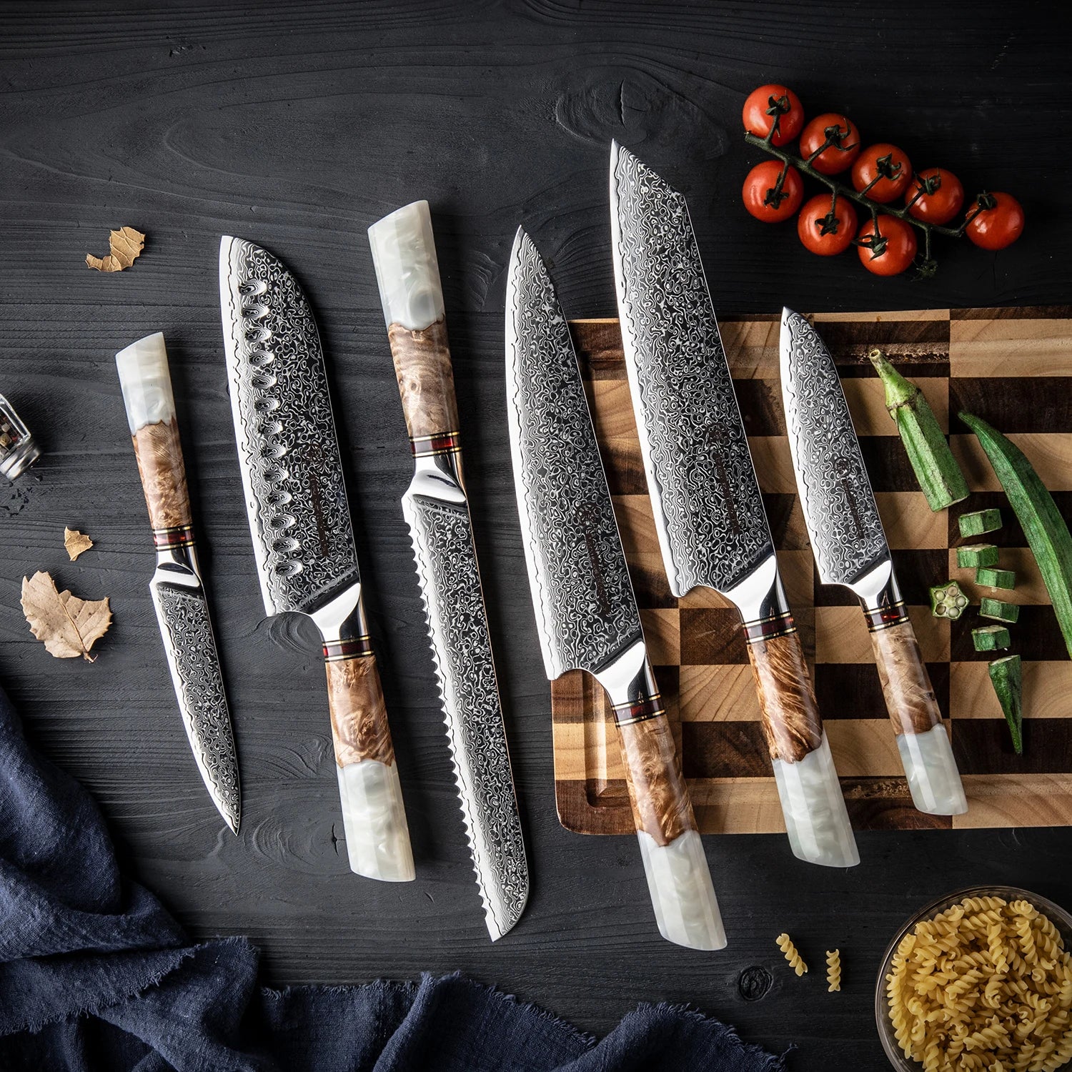 Set of patterned kitchen knives with wood and resin handles on a dark table beside a cutting board with vegetables.