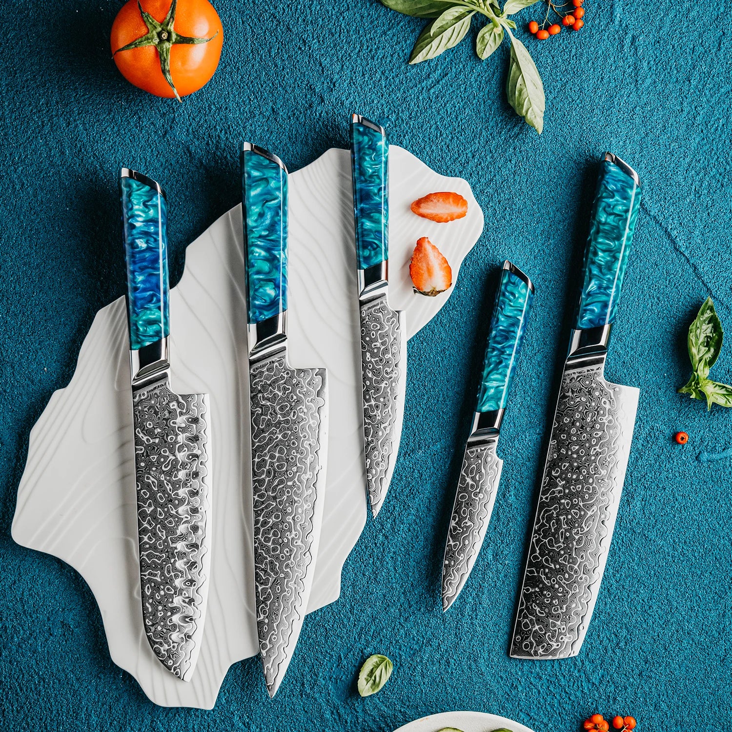 Set of damascus steel kitchen knives with blue resin handles on a textured blue surface, surrounded by herbs and vegetables.